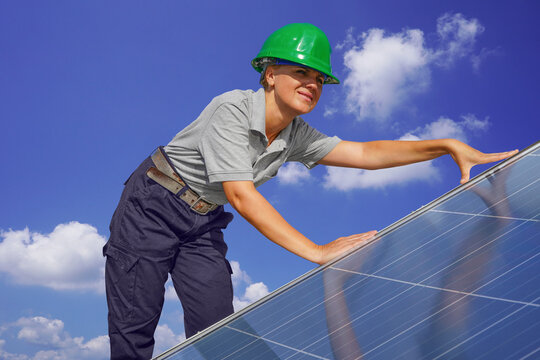 Woman engineer, electrician in green protective helmet against background of cloudy sky installing solar photovoltaic panel system. Professional electrician mounting solar panel. Alternative energy