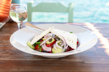 Traditional Greek Salad served in tavern, traditional greece food with Aegean sea as background. Tomatoes, cucumber, onions, olives, peppers, cappers and olive oil