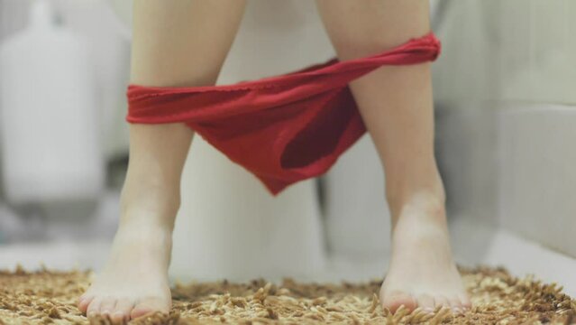 Woman is sitting on the toilet in a home bathroom, taking off her red panties and scratching her leg. Close-up of feet. The concept of constipation and diarrhea.