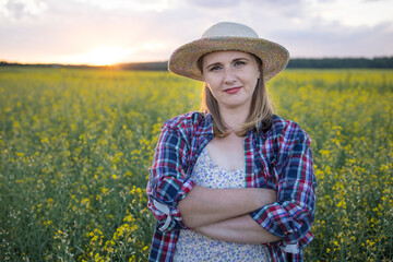 A beautiful middle-aged farmer woman in a straw hat and a plaid shirt stands in a field of flowering rapeseed in the sunlight