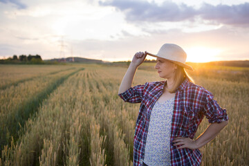 A beautiful middle-aged farmer woman in a straw hat and a plaid shirt stands in a field of golden ripening wheat during the daytime in the sunlight