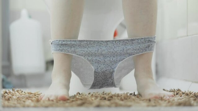 Funny routine. Woman is sitting on the toilet in a home bathroom, takes of two panties and dangles legs. Close-up of feet. The concept of women's health and virginity.