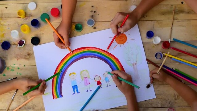 Group of children drawing with colored pencils and paints on wooden floor. Kids make painting family and bright rainbow. Top view.