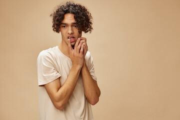 Confused awesome tanned curly man in basic t-shirt touches cheeks posing isolated on over beige pastel background. Fashion New Collection offer. People and Emotions concept. Free place for ad