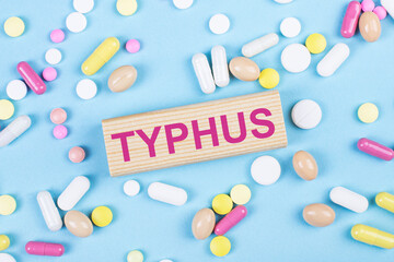 On a blue background, multi-colored pills and a wooden block with the text TYPHUS. View from above. Medical concept