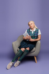 Student in plaid uniform holding digital tablet while sitting on armchair on purple background