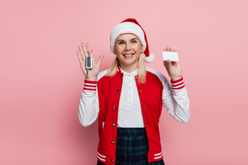 Cheerful blonde woman in santa hat holding driving license and car key on pink background