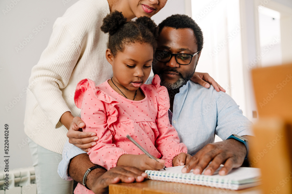 Wall mural African american girl writing in notepad sitting with parents by table