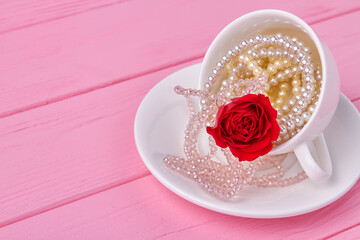 White cup full of pearl necklaces and red flower. Pink wooden desk.