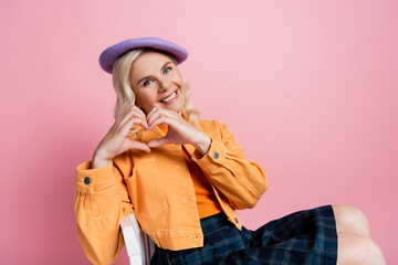 Smiling woman in beret showing heart gesture while sitting on chair isolated on pink