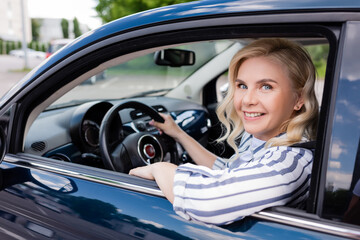 Fototapeta na wymiar Cheerful blonde woman looking at camera while driving auto during course