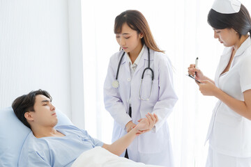 Female doctor and young male patient who lie on the bed while checking pulse, consult and explain with nurse taking note and supporting in hospital wards.