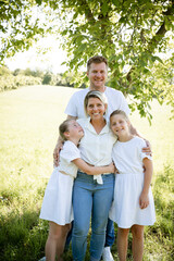Naklejka na ściany i meble beautiful family with two girls is standing in meadow near walnut tree and has light outfit on and jeans and is happy