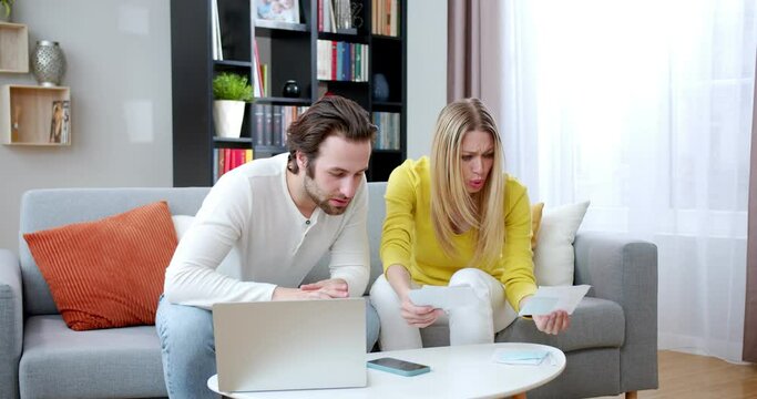 Caucasian young married couple hacing problems with bills and taxes. At home. Man and woman sitting on couch in living room and worrying about lack of money. Stressed and in worryies.