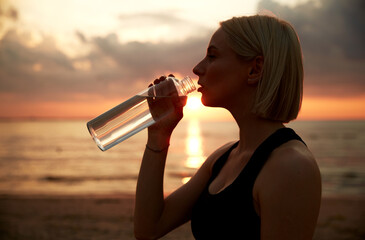 fitness, sport, and healthy lifestyle concept - woman drinking water from bottle on beach over sunset