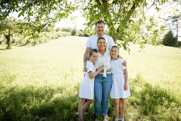 Fototapeta premium beautiful family with two girls is standing in meadow near walnut tree and has light outfit on and jeans and is happy