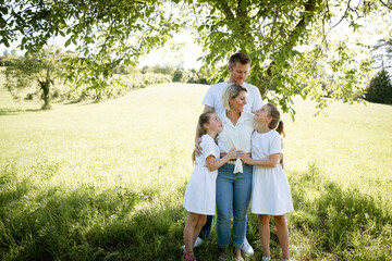 Fototapeta premium beautiful family with two girls is standing in meadow near walnut tree and has light outfit on and jeans and is happy