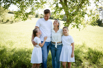 Fototapeta premium beautiful family with two girls is standing in meadow near walnut tree and has light outfit on and jeans and is happy