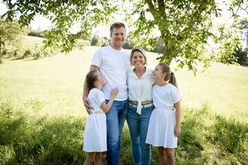 beautiful family with two girls is standing in meadow near walnut tree and has light outfit on and jeans and is happy