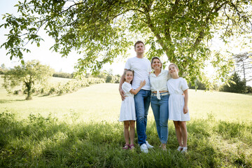 Fototapeta premium beautiful family with two girls is standing in meadow near walnut tree and has light outfit on and jeans and is happy