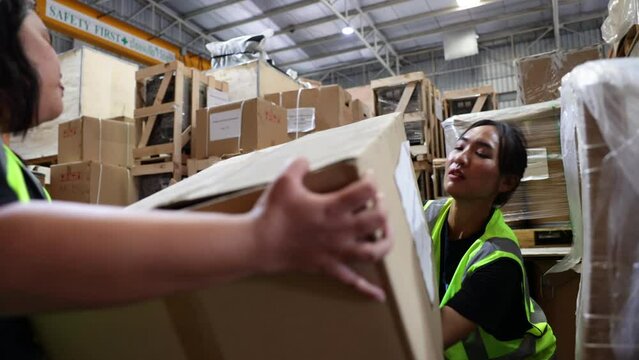 Two women worker carrying and lifting heavy cardboard box. Asian obese female warehouse worker in safety vest and hardhat helmet working in warehouse factory industrial.