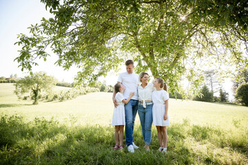 beautiful family with two girls is standing in meadow near walnut tree and has light outfit on and jeans and is happy