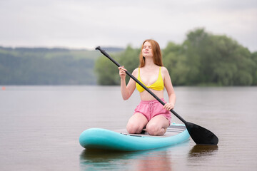 A beautiful redheaded girl is sailing on a sapboard on the river. Young woman surfing at sunrise.