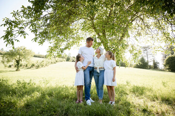 beautiful family with two girls is standing in meadow near walnut tree and has light outfit on and jeans and is happy