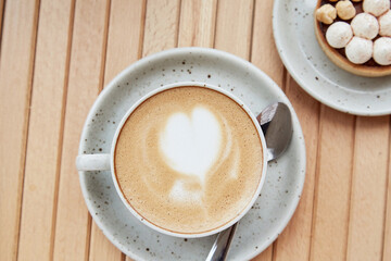Heart shaped foam of cappuccino in white ceramic cup and hazelnut tart on wooden table at the terrace. Aesthetic breakfast top view. Flat lay food