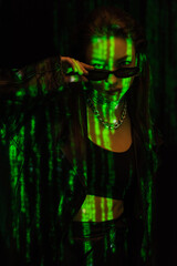 young woman in silver necklaces holding dark sunglasses and looking at camera in green neon light isolated on black