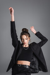 trendy woman in crop top and black jacket standing with arms up isolated on grey