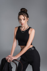Fototapeta na wymiar trendy woman in black crop top and silver necklace posing near chair isolated on grey