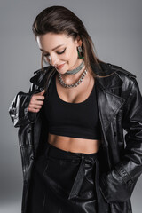 pretty woman in black leather clothes and silver necklaces smiling isolated on grey