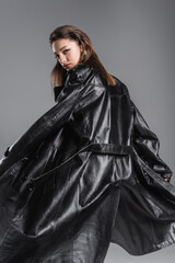 young and stylish woman in black leather coat looking at camera while posing isolated on grey