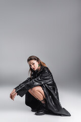 Naklejka premium fashionable brunette woman in black leather outfit sitting on haunches and looking at camera on grey background