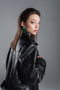 brunette woman with makeup standing in black leather coat and looking at camera isolated on grey