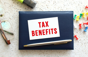 Text TAX BENEFITS on a business card lying on a blue notebook next to the glasses and stationery.