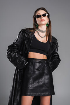 young woman in dark sunglasses and black leather clothing posing with hands in pockets isolated on grey