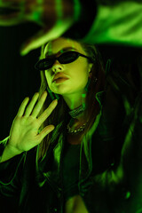young woman in dark sunglasses gesturing in green neon light isolated on black
