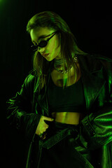 trendy woman in sunglasses and crop top standing with hand on hip in green light isolated on black