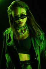 stylish woman in crop top and dark sunglasses looking at camera in green light isolated on black