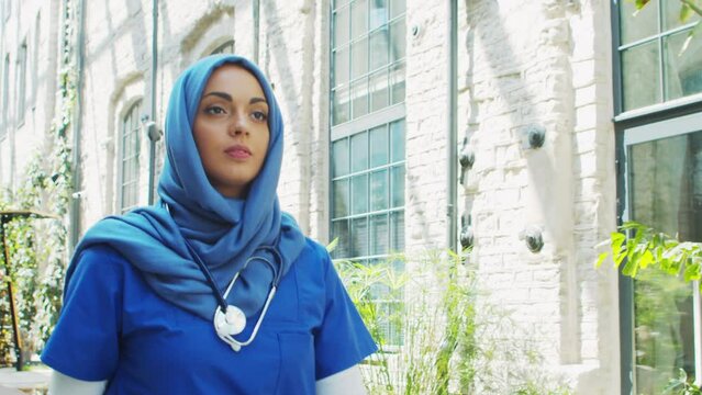 Professional Muslim medical doctor works in front of hospital office. Portrait of young and attractive Middle Eastern female physician in hijab.