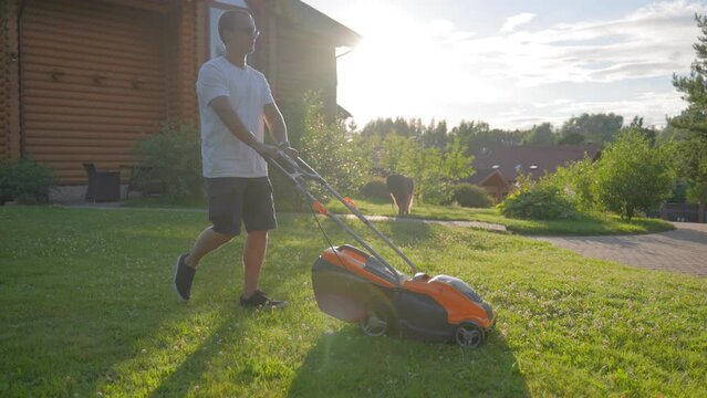 Middle-aged man enjoys taking care of big yard lawn. Mature gardener wear white t-shirt and glasses cuts grass with orange mover smiling at back sunset light