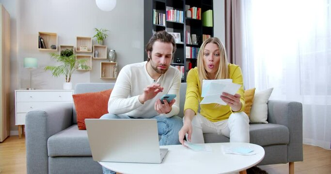 Stressed upset man calculating with smartphone taves and bills while woman showing checks and numbers. At home. Young family having problems with money. Wife and husband getting invoices.