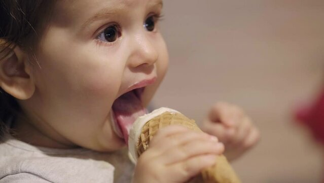 Closeup portrait of cute caucasian baby girl eating licking ice cream in waffle cone. Face and mouth smeared, smeared with cream.