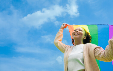 Naklejka premium lgbtq concept. Positive African girl with afro curly hair holding rainbow flag isolated in studio.An African American young woman holding and raising a rainbow flag over the blue sky.
