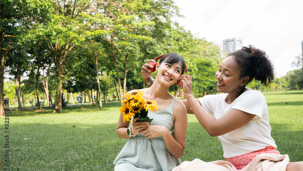 Wall mural Smiling two young diversity African American Asian women friends having fun in a nature, park queer non-binary gender identity, gay lesbian love romance, boho summer vacation style.Listening music.