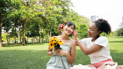 Fototapeta premium Smiling two young diversity African American Asian women friends having fun in a nature, park queer non-binary gender identity, gay lesbian love romance, boho summer vacation style.Listening music.
