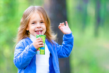 Cheerful little girl is drinking juice outdoors. Walk in the woods, summer sunny day