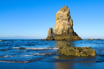Fototapeta na wymiar Waves gently surround The Needles sea stack at Cannon Beach Oregon with a perfect morning blue sky